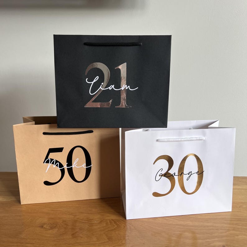 Puede incluir: Tres bolsas de regalo en negro, beige y blanco, cada una con un n&uacute;mero y un nombre. La bolsa negra tiene '21' y 'Liam' en oro rosa. La bolsa beige muestra '50' y 'Mike' en negro. La bolsa blanca muestra '30' y 'George' en dorado. Todas las bolsas tienen asas.
