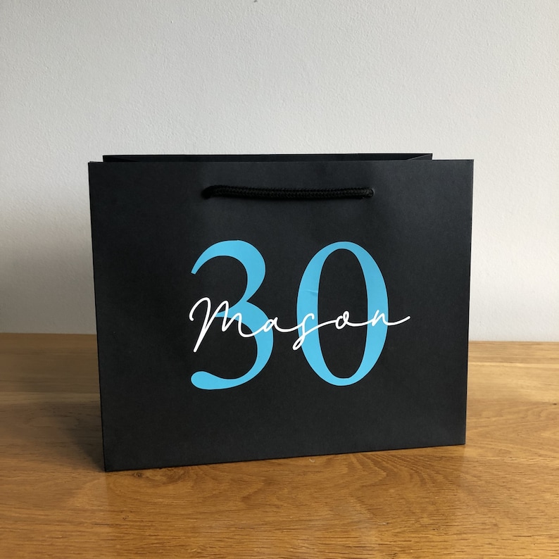 Puede incluir: Bolsa de regalo negra con asa de cuerda negra. La bolsa muestra el n&uacute;mero "30" en azul claro, con el nombre "Mason" en cursiva debajo. La bolsa est&aacute; sobre una superficie de madera.