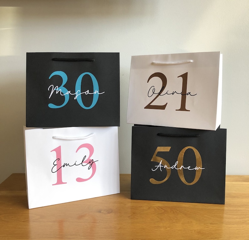 Puede incluir: Cuatro bolsas de regalo en blanco y negro, cada una con un n&uacute;mero y un nombre. Los n&uacute;meros son 30, 21, 13 y 50, en azul, marr&oacute;n, rosa y dorado. Los nombres son Mason, Olivia, Emily y Andrew, en cursiva.