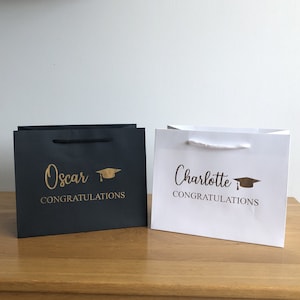 May include: Two graduation gift bags, one black and one white, with gold lettering and a graduation cap icon. The black bag says "Oscar Congratulations" and the white bag says "Charlotte Congratulations".