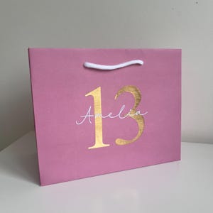 Puede incluir: Una bolsa de regalo rosa con un asa de cuerda blanca. El n&uacute;mero "13" est&aacute; impreso en dorado, con el nombre "Amelia" en cursiva debajo. La bolsa est&aacute; sobre una superficie blanca.