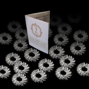 May include: A collection of silver massage rings scattered around a folded white card. The card features a gold emblem with the letter "Z" and the text "BAGUA ZEN MASSAGE RING". The rings have a spiky texture.
