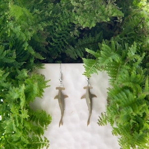 May include: A pair of dangle earrings featuring a hammerhead shark design. The earrings are a muted olive green color and hang from silver-colored hooks. The earrings are displayed against a backdrop of lush green foliage.