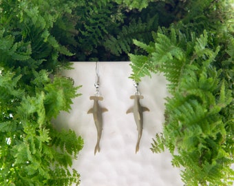 May include: A pair of dangle earrings featuring a hammerhead shark design. The earrings are a muted olive green color and hang from silver-colored hooks. The earrings are displayed against a backdrop of lush green foliage.