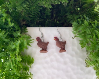 May include: A pair of brown chicken earrings with silver hooks. The chickens are standing on a small base.