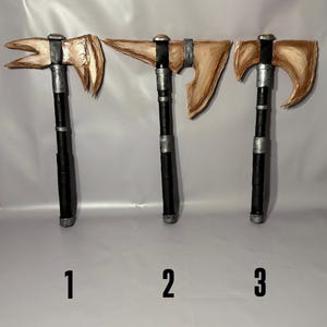 May include: Three handcrafted axes with wooden blades and black handles are displayed against a white backdrop. The blades have a light brown finish, while the handles are wrapped in black material. Each axe has a unique blade shape, and numbers 1, 2, and 3 are below them.