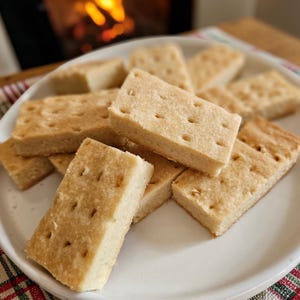 May include: A plate of golden-brown shortbread cookies. Each rectangular cookie has a slightly rough texture and small, evenly spaced holes. The cookies are stacked on a white plate, with a warm, blurred background.