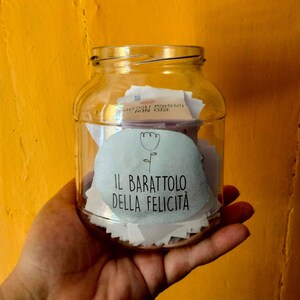May include: A clear glass jar filled with small pieces of paper, with a white label that says "IL BARATTOLO DELLA FELICIT&Agrave;" and a small flower illustration. The jar is being held in a hand against a yellow background.