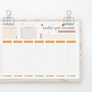 May include: Weekly planner with the Italian text "Goditi ogni momento" and "Calendario Settimanale". The planner has sections for each day of the week, with time slots and space for notes. It is held in place by silver binder clips.