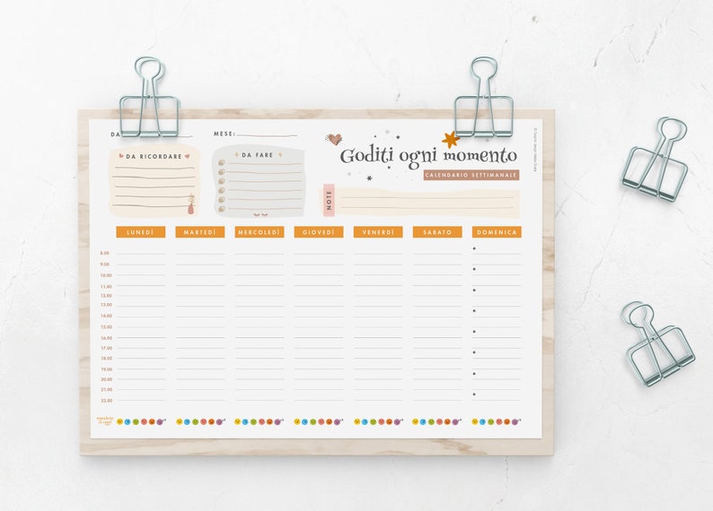 May include: Weekly planner with the Italian text "Goditi ogni momento" and "Calendario Settimanale". The planner has sections for each day of the week, with time slots and space for notes. It is held in place by silver binder clips.