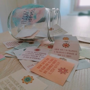 Puede incluir: Un tarro de vidrio lleno de peque&ntilde;as tarjetas coloridas con mensajes inspiradores escritos en italiano. El tarro est&aacute; volcado y las tarjetas est&aacute;n esparcidas sobre una superficie de madera clara. Las tarjetas tienen una variedad de dise&ntilde;os y colores, incluyendo rosa, azul, verde y amarillo. El texto en las tarjetas est&aacute; en italiano e incluye frases como "Cadere non &egrave; un fallimento" y "Ci sono due modi di affrontare le difficolt&agrave;".