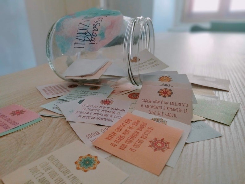 Puede incluir: Un tarro de vidrio lleno de peque&ntilde;as tarjetas coloridas con mensajes inspiradores escritos en italiano. El tarro est&aacute; volcado y las tarjetas est&aacute;n esparcidas sobre una superficie de madera clara. Las tarjetas tienen una variedad de dise&ntilde;os y colores, incluyendo rosa, azul, verde y amarillo. El texto en las tarjetas est&aacute; en italiano e incluye frases como "Cadere non &egrave; un fallimento" y "Ci sono due modi di affrontare le difficolt&agrave;".