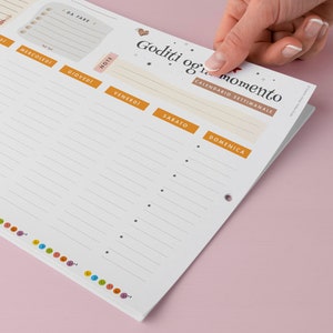 May include: A weekly planner with a white background and orange and beige accents. The planner has sections for each day of the week, notes, and a to-do list. The top of the planner has the text "Goditi ogni momento" and "Calendario Settimanale".