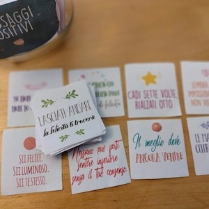 May include: A jar filled with small cards containing positive messages in Italian. The cards are on a wooden surface. The jar has a black label that says "Messaggi Positivi" which translates to "Positive Messages".