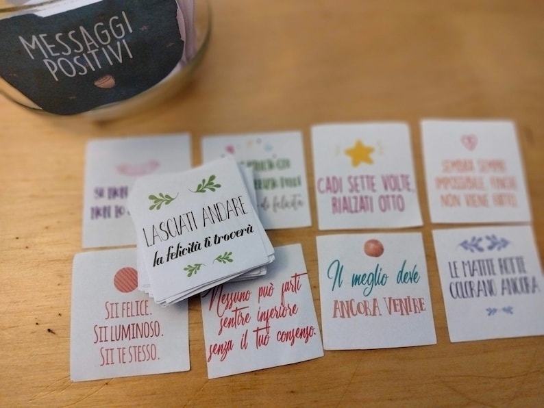 May include: A jar filled with small cards containing positive messages in Italian. The cards are on a wooden surface. The jar has a black label that says "Messaggi Positivi" which translates to "Positive Messages".