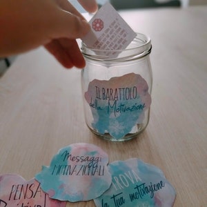 Puede incluir: Un tarro de vidrio con una etiqueta que dice "Il Barattolo della Motivazione" (El tarro de la motivaci&oacute;n). El tarro est&aacute; lleno de peque&ntilde;os trozos de papel con mensajes motivacionales. Hay cuatro tarjetas de papel de colores con mensajes motivacionales: "Pensa Positivo!" (&iexcl;Piensa positivo!), "Messaggi Motivazionali" (Mensajes motivacionales), "Trova la tua motivazione" (Encuentra tu motivaci&oacute;n).