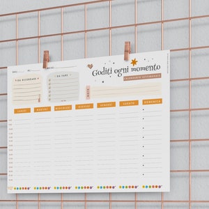 May include: A weekly planner with the text "Goditi ogni momento" is clipped to a rose gold wire grid. The planner has sections for notes, to-do lists, and a daily schedule. The grid is mounted on a light gray wall.