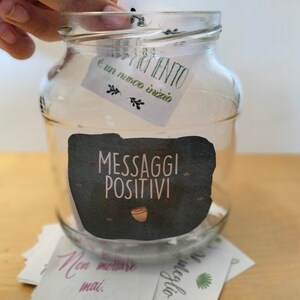 May include: A clear glass jar with a black and white label that says "Messaggi Positivi" and a small pink flower graphic. The jar is filled with small white slips of paper. A hand is placing a white slip of paper with the text "Ogni momento &egrave; un nuovo inizio" into the jar.