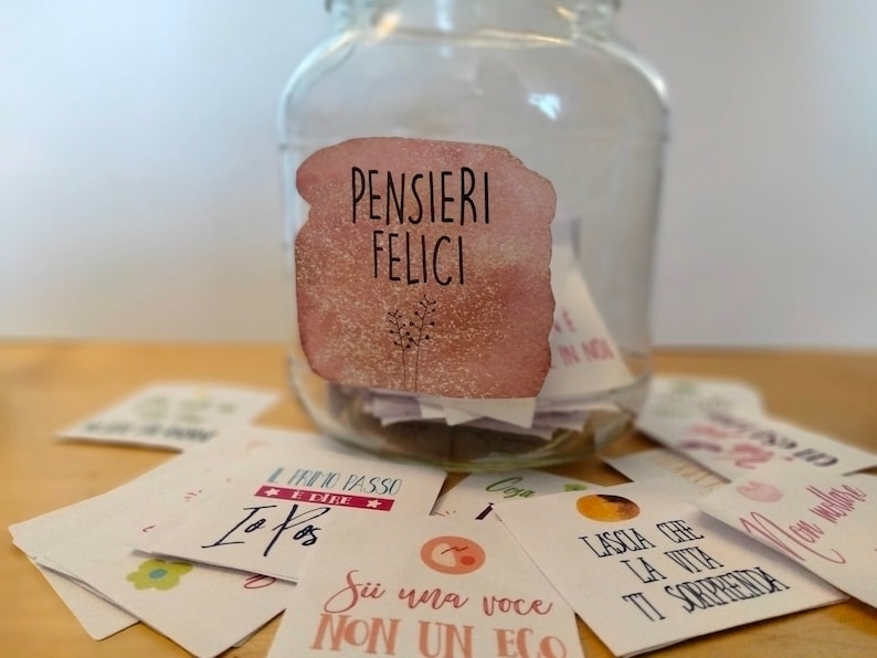May include: A glass jar with a pink and white watercolor label that says "PENSIERI FELICI" filled with small white cards. The cards have inspirational messages written on them in Italian.