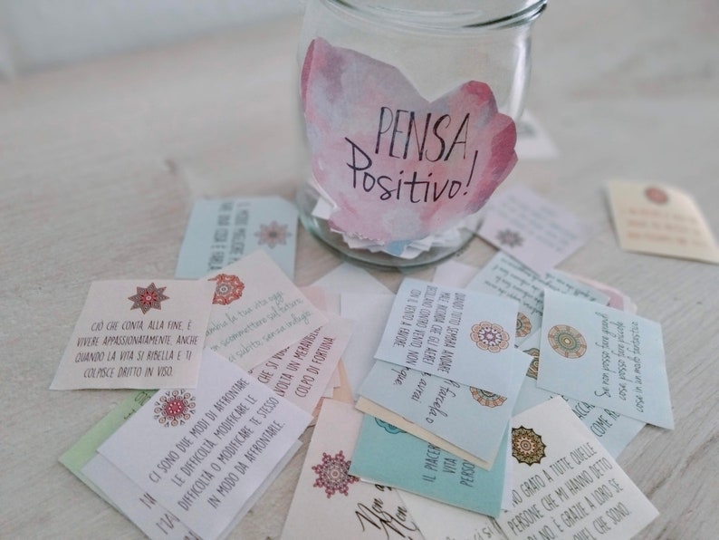 Puede incluir: Un tarro de vidrio con un dise&ntilde;o de acuarela rosa y blanco que dice "PENSA Positivo!" El tarro est&aacute; lleno de peque&ntilde;as tarjetas con mensajes inspiradores escritos en italiano.