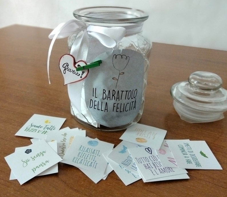 May include: A glass jar with a white ribbon and a label that reads "Il Barattolo Della Felicita" (The Jar of Happiness). The jar is filled with small cards with inspirational messages written in Italian. The jar is sitting on a wooden surface.