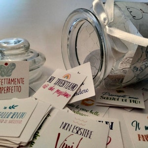 May include: A glass jar with a white ribbon and a white label with the text "IL BARATTOLO DELLA FELICITA" on it. The jar is filled with small cards with inspirational quotes in Italian. The quotes include "PERFETTAMENTE IMPERFETTO", "COM FALLIMENTO E UN PASSO VERSO IL SUCCESSO", "MI DIVENTIAMO CIO CHE PENSIAMO", "NON ESISTERE", "PREOCCUPATI MENO. SORRIDI DI PIU!" and "IL MEGLIO DE ANCORA VIENE".