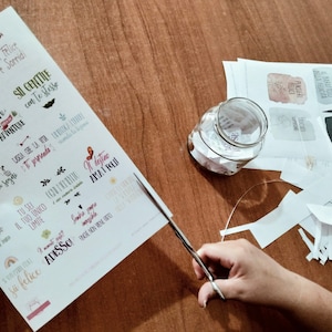 May include: A sheet of paper with motivational quotes printed in Italian. The quotes are printed in a variety of fonts and colors. The paper is being cut with scissors. There is a glass jar filled with small pieces of paper on a wooden table.