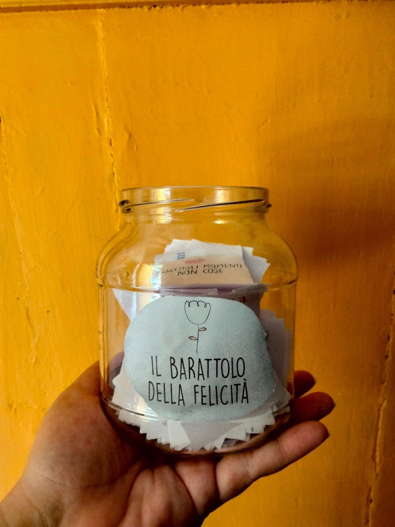 May include: A clear glass jar filled with small pieces of paper, with a white label that says "IL BARATTOLO DELLA FELICIT&Agrave;" and a small flower illustration. The jar is being held in a hand against a yellow background.