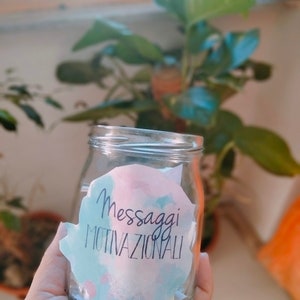 Puede incluir: Un frasco de vidrio lleno de peque&ntilde;os papeles con mensajes motivacionales escritos en ellos. El frasco tiene una pegatina de acuarela rosa, azul y blanca que dice "Messaggi Motivazionali".