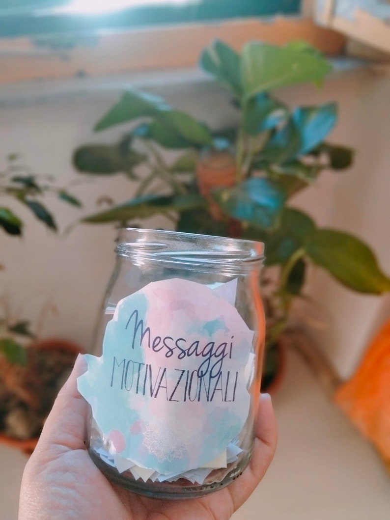 Puede incluir: Un frasco de vidrio lleno de peque&ntilde;os papeles con mensajes motivacionales escritos en ellos. El frasco tiene una pegatina de acuarela rosa, azul y blanca que dice "Messaggi Motivazionali".