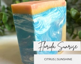 May include: A rectangular bar of soap featuring a layered design with a peach and yellow top section, a blue and white marbled middle, and a solid yellow base. The text Florida Sunrise and CITRUS | SUNSHINE is printed on the front.