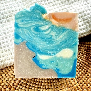 Puede incluir: Una pastilla de jab&oacute;n hecha a mano con un dise&ntilde;o en capas. El jab&oacute;n presenta tonos de azul, blanco y beige, que se asemejan a una escena costera. El jab&oacute;n descansa sobre una estera tejida y una toalla blanca texturizada.