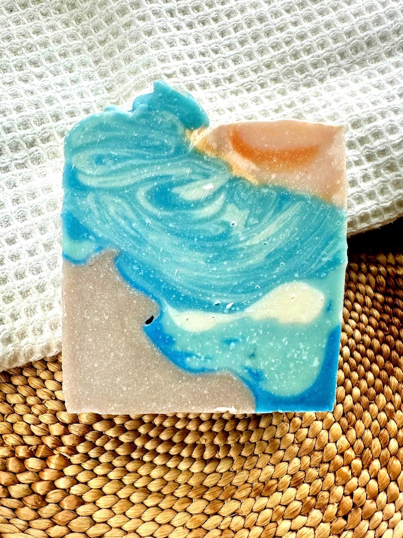 Puede incluir: Una pastilla de jab&oacute;n hecha a mano con un dise&ntilde;o en capas. El jab&oacute;n presenta tonos de azul, blanco y beige, que se asemejan a una escena costera. El jab&oacute;n descansa sobre una estera tejida y una toalla blanca texturizada.