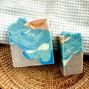 Puede incluir: Dos jabones artesanales con tem&aacute;tica de playa. Los jabones presentan capas de colores azul, blanco y beige, que se asemejan a las olas del oc&eacute;ano y la arena. Los jabones est&aacute;n sobre una cesta tejida y una toalla blanca.