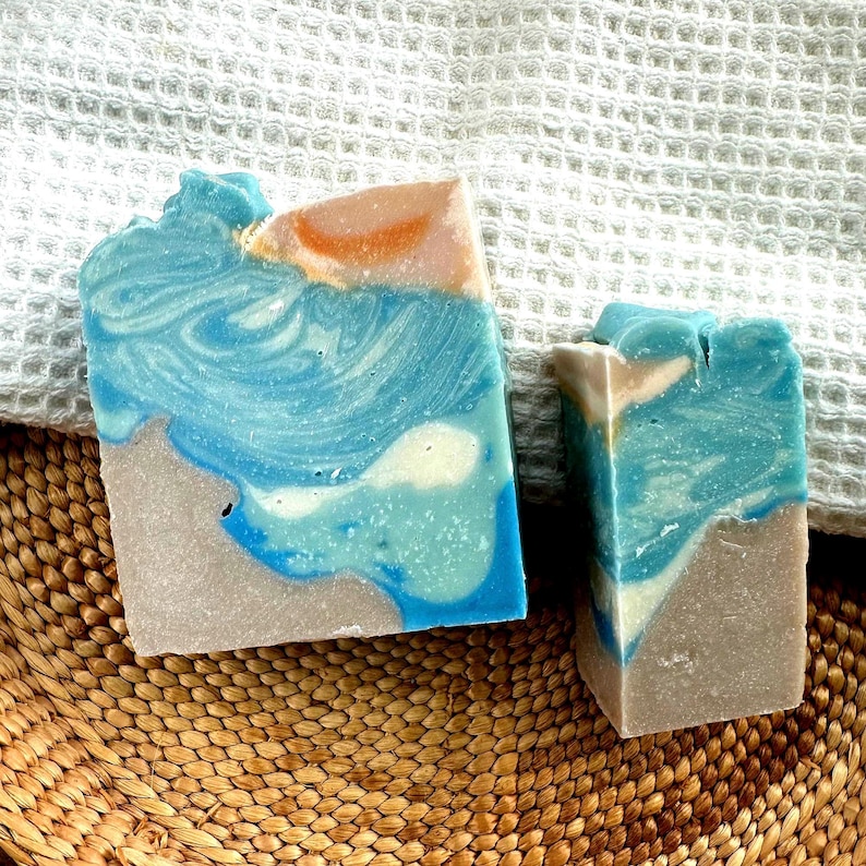 Puede incluir: Dos jabones artesanales con tem&aacute;tica de playa. Los jabones presentan capas de colores azul, blanco y beige, que se asemejan a las olas del oc&eacute;ano y la arena. Los jabones est&aacute;n sobre una cesta tejida y una toalla blanca.