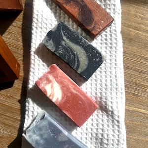 Puede incluir: Cuatro barras rectangulares de jabón artesanal en varios colores y patrones. La barra superior es marrón con remolinos oscuros, la segunda es negra y gris, la tercera es roja y blanca, y la inferior es azul y gris. Los jabones están sobre una toalla blanca texturizada.
