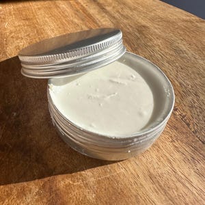 Op de afbeelding: Een kleine, ronde, zilverkleurige metalen container met een schroefdeksel, gedeeltelijk geopend. De container bevat een witte, romige substantie. De container staat op een houten oppervlak, met het deksel ernaast.