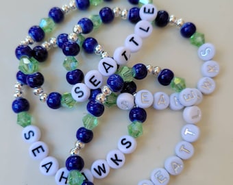 May include: Three beaded bracelets with alternating blue and green beads and white beads with the words "SEATTLE" and "HAWKS" spelled out.