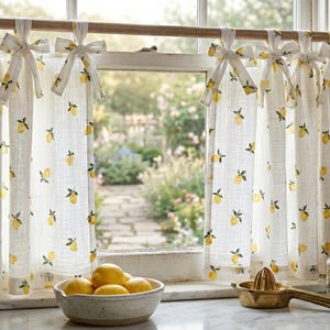 May include: A pair of white cafe curtains featuring a repeating yellow lemon print with green leaves. The curtains are hung on a wooden rod using fabric ties at the top, creating a gathered effect. The fabric has a visible textured weave.