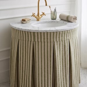Op de afbeelding: Een badkamermeubel met een wit marmeren blad, een halfronde wastafel en een messing kraan. De onderkant is bedekt met een geplooide stoffen rok met verticale olijfgroene en beige strepen. Een zeepbakje, tandenborstelhouder en handdoek staan bovenop.