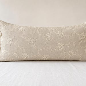 Peut inclure: Un coussin lombaire rectangulaire de couleur beige clair orné d'une superposition de dentelle florale blanche complexe. Le côté du coussin est fixé par trois liens en tissu beige assortis, créant un détail noué décoratif sur le bord.