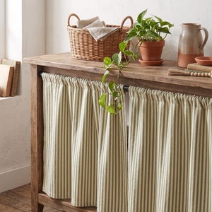May include: A pair of cafe curtains featuring a vertical striped pattern in muted sage green and off-white. The fabric is gathered along a thin black metal rod, which is mounted to the front of a wooden console table with a natural finish.