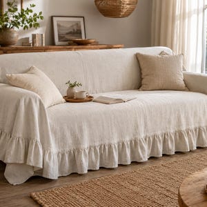 May include: A beige linen sofa cover featuring a ruffled skirt along the bottom edge. The cover is draped over a sofa and includes two matching square throw pillows placed on the seat cushions, providing a textured and neutral fabric appearance.