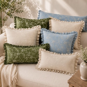 May include: A collection of rectangular throw pillows featuring a delicate white floral embroidery pattern on solid backgrounds of sage green, light blue, and cream. Each pillow is finished with a ruffled fabric border along the edges.