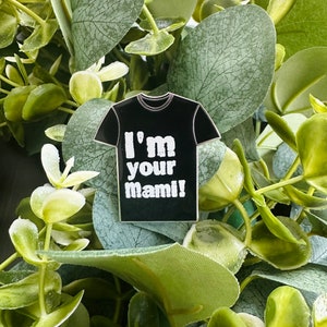 Puede incluir: Un pin de esmalte negro con forma de camiseta con el texto "I'm your mami!" en blanco.