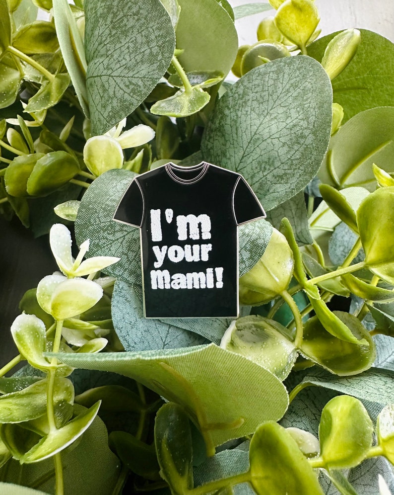 Puede incluir: Un pin de esmalte negro con forma de camiseta con el texto "I'm your mami!" en blanco.