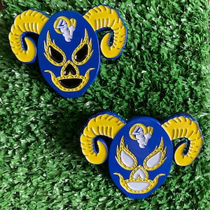 May include: Two blue and yellow wrestling masks with horns. The masks have white eyes and a white mouth with teeth. The masks are on a green background.