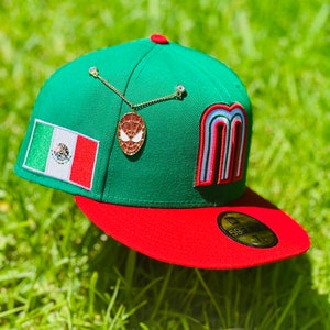 Puede incluir: Una gorra de béisbol verde y roja con un parche de la bandera mexicana, una cadena dorada con un colgante de Spider-Man rojo y blanco, y una letra M arcoíris en el lateral. La gorra tiene un logotipo de New Era en la parte posterior.