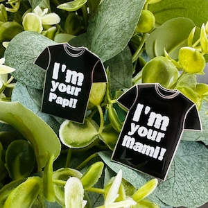 Puede incluir: Dos pines de esmalte negro con forma de camiseta con texto blanco. El primer pin dice "I'm your Papi!" y el segundo pin dice "I'm your mami!"