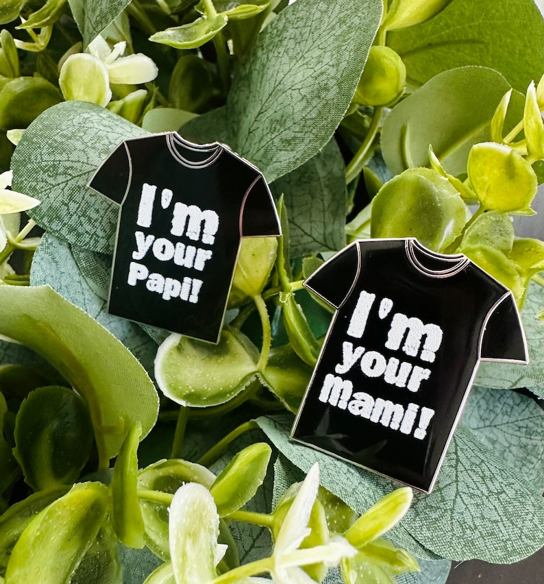 Puede incluir: Dos pines de esmalte negro con forma de camiseta con texto blanco. El primer pin dice "I'm your Papi!" y el segundo pin dice "I'm your mami!"